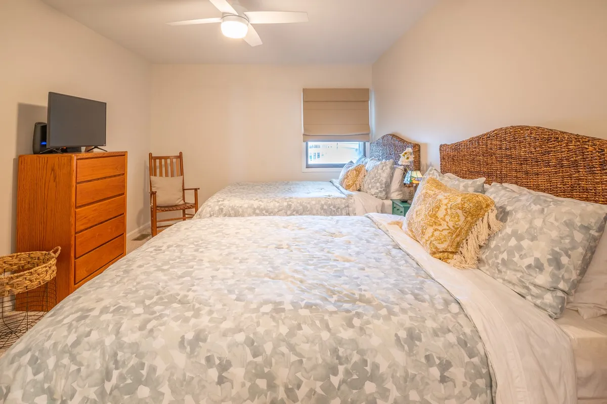 A guest bedroom with two comfortable beds adorned with decorative pillows, a wooden dresser, and an inviting rocking chair placed in the corner. Soft morning light brightens the space, creating a welcoming environment for families or friends.