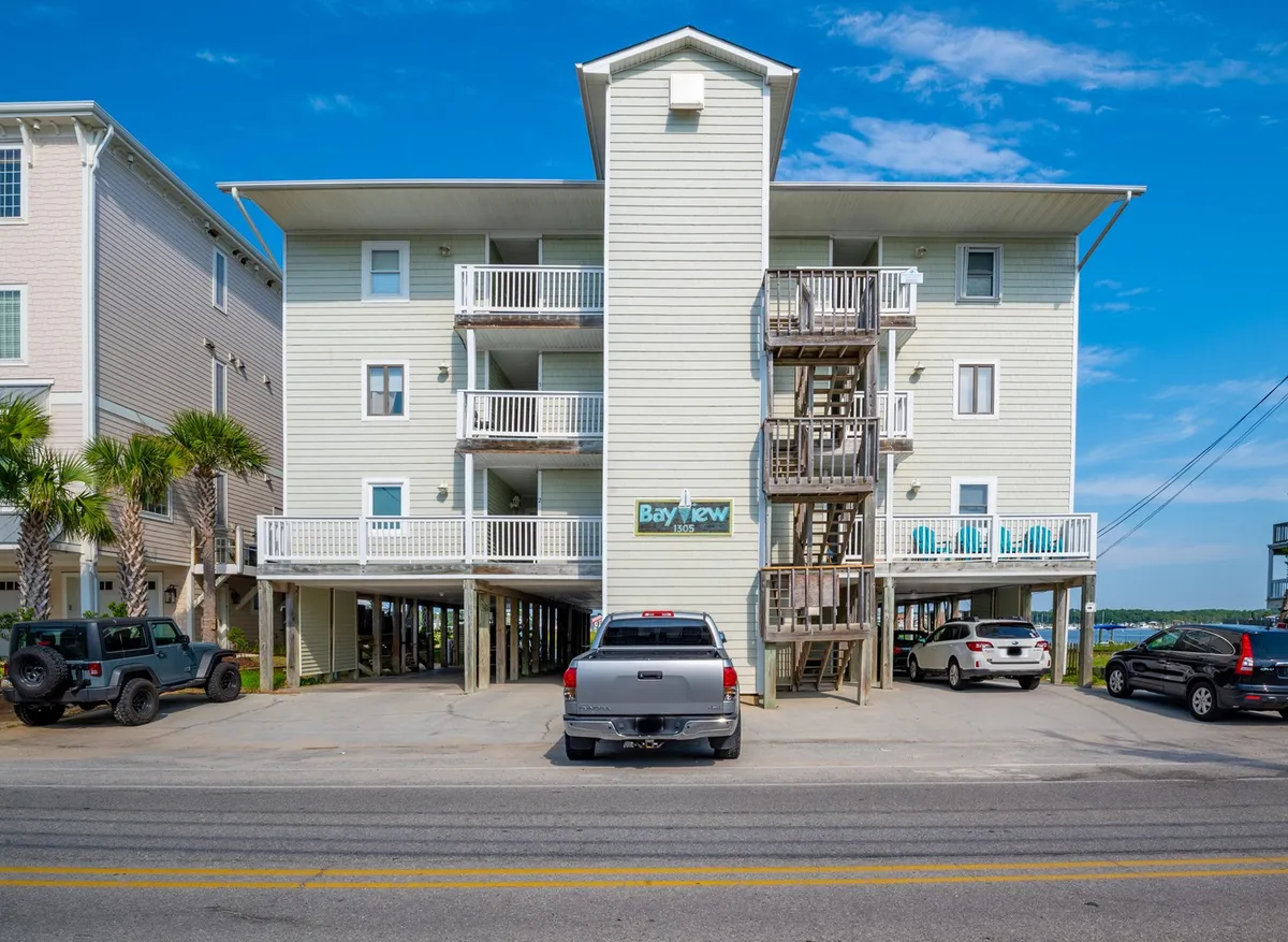 The image shows the inviting exterior of the Bayview Bliss condo building at Carolina Beach, with the prominent 'Bayview' sign, palm trees swaying, balconies overlooking the canal, and a relaxed beach-town atmosphere. Parked vehicles suggest easy access for guests, and the stairwell leads up to private porches ideal for morning coffee or sunset views.