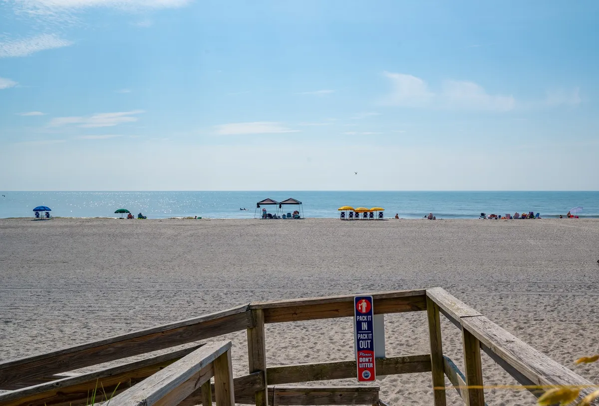 A lively, sun-soaked beach scene featuring the ocean waves, colorful umbrellas and cabana tents, with the boardwalk visible in the background. People are enjoying classic beach activities like sunbathing, swimming, and playing games, all set against an inviting coastal backdrop.
