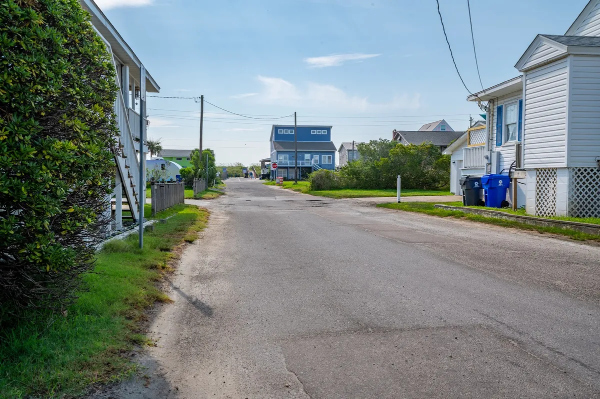 This exterior image shows a quiet residential street lined with houses, greenery, and utility poles. The atmosphere is peaceful, suggesting a secure and comfortable neighborhood setting. The street view also hints at the accessibility of parking and the everyday convenience described in the section.