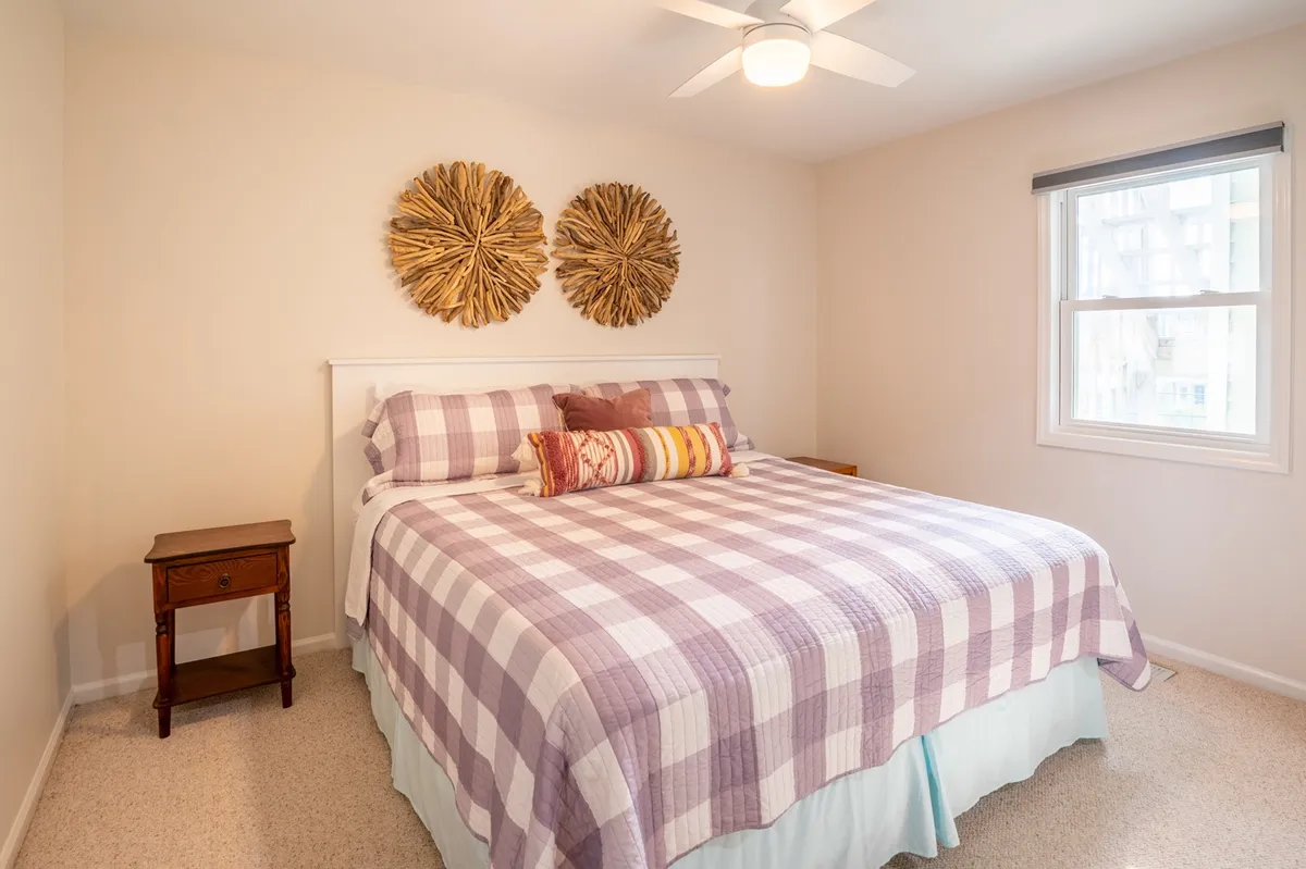 A cozy bedroom featuring a bed with plaid bedding, decorative wall art above the headboard, a nightstand with a lamp, and natural light filtering through the window. The room exudes warmth and relaxation—ideal for unwinding at night.