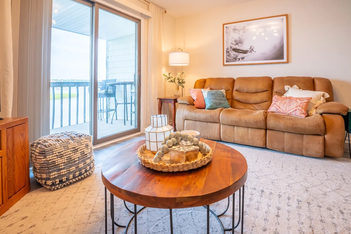 A charming living space with a brown couch and wooden coffee table set beside a large window that frames the tranquil waterfront. The cozy furnishings invite relaxation, reading, or enjoying quiet mornings while basking in natural light and the changing colors of the sky at sunset.