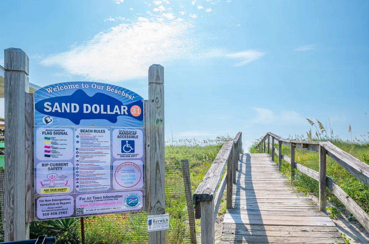 This image features a welcoming exterior scene with a prominent welcome sign, a wooden walkway surrounded by beach grass, and a clear path leading toward the beach. The relaxed and inviting mood sets the tone for seamless arrivals, suggesting both convenience and close proximity to the ocean.
